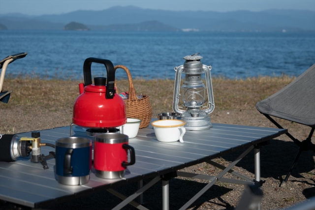 海辺のキャンプテーブルに赤いケトルとランタンを並べた朝の風景